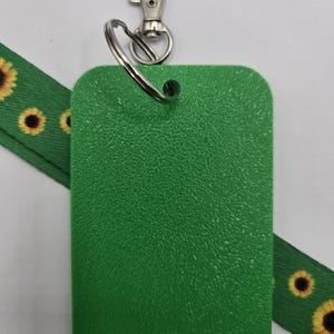 Puede incluir: Un porta credenciales rectangular de pl&aacute;stico verde texturizado unido a una argolla de metal plateado y un mosquet&oacute;n. Una cinta de tela verde con un patr&oacute;n repetitivo de girasoles de p&eacute;talos amarillos y centros oscuros pasa por detr&aacute;s.
