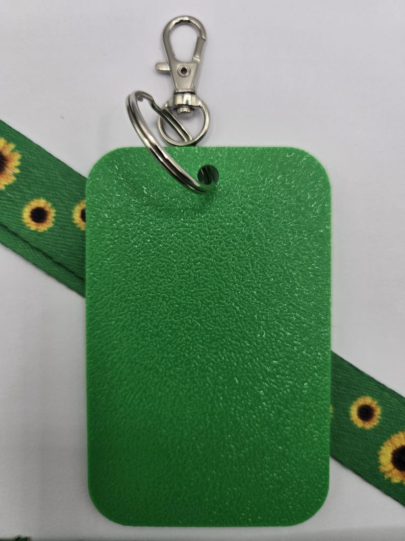 Puede incluir: Un porta credenciales rectangular de pl&aacute;stico verde texturizado unido a una argolla de metal plateado y un mosquet&oacute;n. Una cinta de tela verde con un patr&oacute;n repetitivo de girasoles de p&eacute;talos amarillos y centros oscuros pasa por detr&aacute;s.