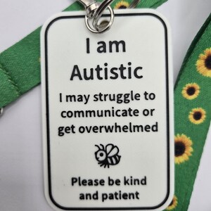 Puede incluir: Una etiqueta rectangular blanca con texto negro que dice I am Autistic, I may struggle to communicate or get overwhelmed y Please be kind and patient, con un icono de abeja. Est&aacute; sujeta a un cord&oacute;n verde con un patr&oacute;n de girasoles.