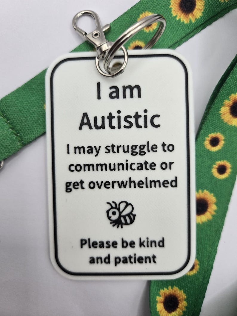 Puede incluir: Una etiqueta rectangular blanca con texto negro que dice I am Autistic, I may struggle to communicate or get overwhelmed y Please be kind and patient, con un icono de abeja. Est&aacute; sujeta a un cord&oacute;n verde con un patr&oacute;n de girasoles.