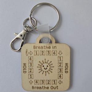 May include: Wooden square pendant keychain with a breathing exercise guide. The pendant has a sun illustration and the words "Breathe In" and "Breathe Out." It includes a silver clasp and ring. The pendant is approximately 4 cm square.