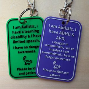 May include: Two keychains with silver clasps. One is green and the other is purple. The green keychain reads, "I am Autistic, I have a learning disability & I have limited speech. I have no danger awareness." The purple keychain reads, "I am Autistic, I have ADHD & APD. I struggle to communicate, I act on impulse & I get overwhelmed. I have no danger awareness."