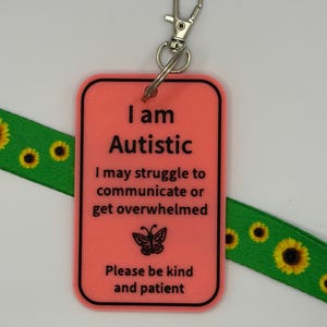 Puede incluir: Una etiqueta rectangular de pl&aacute;stico color coral con borde negro y texto negro que dice: I am Autistic, I may struggle to communicate or get overwhelmed, Please be kind and patient, con un icono de mariposa y un cierre de metal.