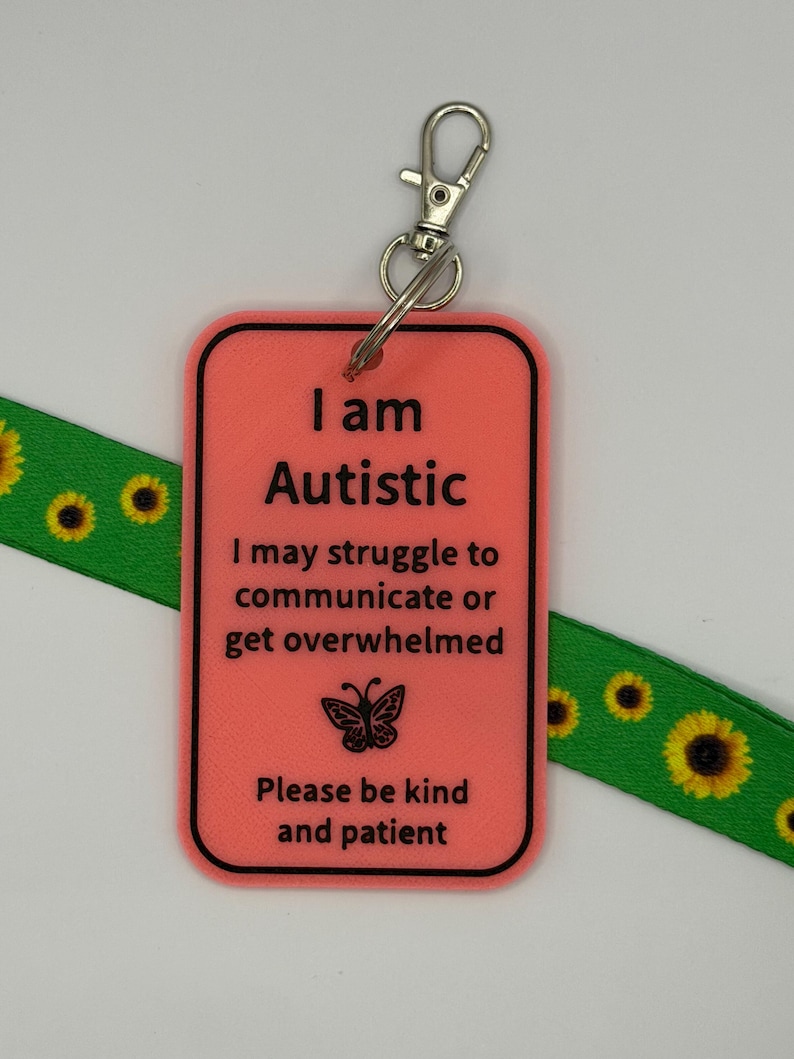 Puede incluir: Una etiqueta rectangular de pl&aacute;stico color coral con borde negro y texto negro que dice: I am Autistic, I may struggle to communicate or get overwhelmed, Please be kind and patient, con un icono de mariposa y un cierre de metal.