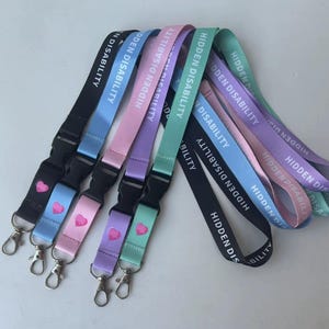 Puede incluir: Una colecci&oacute;n de cinco cordones en negro, azul claro, rosa, morado y verde menta. Cada correa de tela presenta el texto blanco HIDDEN DISABILITY y un peque&ntilde;o icono de coraz&oacute;n rosa sobre una hebilla de pl&aacute;stico negro y un gancho giratorio.