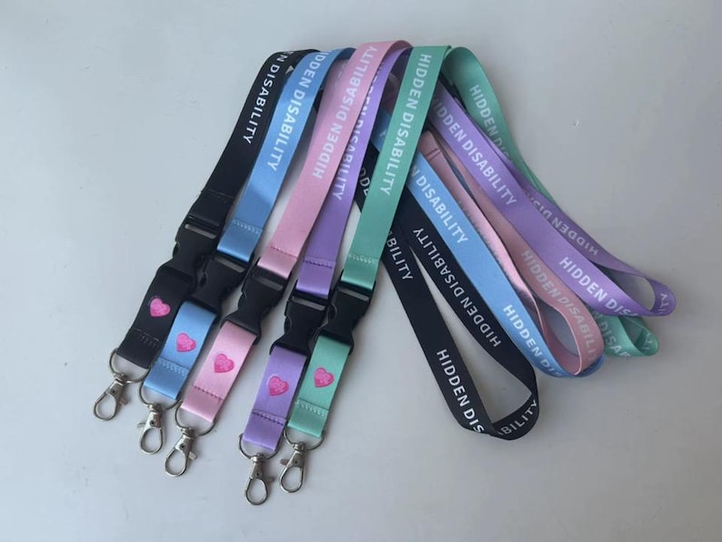 Puede incluir: Una colecci&oacute;n de cinco cordones en negro, azul claro, rosa, morado y verde menta. Cada correa de tela presenta el texto blanco HIDDEN DISABILITY y un peque&ntilde;o icono de coraz&oacute;n rosa sobre una hebilla de pl&aacute;stico negro y un gancho giratorio.