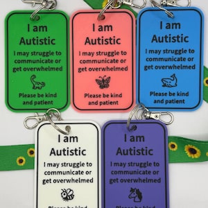 May include: Five rectangular keychains in green, coral, blue, white, and purple. Each keychain has the text "I am Autistic" and a message about communication. The keychains are attached to silver clasps and a sunflower lanyard.