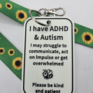 May include: A white rectangular tag with black text reading I have ADHD & Autism, I may struggle to communicate, act on impulse or get overwhelmed, Please be kind and patient, featuring a small bee icon, attached to a green sunflower lanyard.