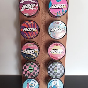 Peut inclure: Un présentoir en bois avec douze boîtes métalliques rondes. Chaque boîte a un couvercle coloré avec le mot "HOLY" et le texte "Boost Your Focus with NewCaff & Vitamins". Les couvercles présentent divers motifs.