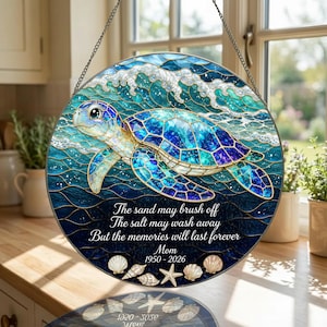 May include: A circular stained glass suncatcher featuring a sea turtle swimming in blue ocean waves. The text reads: The sand may brush off, The salt may wash away, But the memories will last forever, Mom, 1950-2026. Seashells are at the bottom.