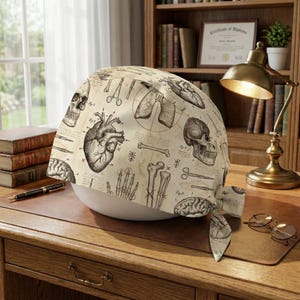 May include: A beige surgical scrub cap featuring anatomical illustrations of the heart, lungs, brain, and bones. The medical-themed cap includes images of surgical tools and a skull. The cap is tied at the back.