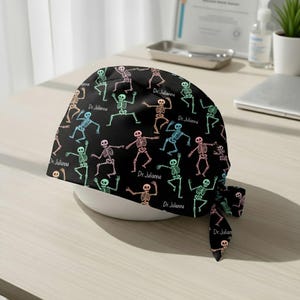 May include: Black surgical scrub cap with a repeating pattern of colorful skeletons in various poses. The text "Dr. Julianna" is printed on the cap. The cap is tied at the back.