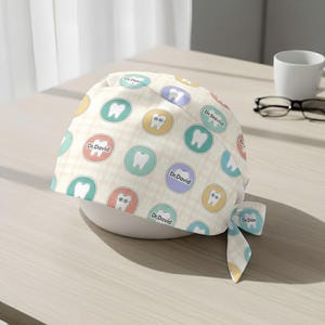 May include: A light beige dental scrub cap with a repeating pattern of colorful circles featuring teeth and the text "Dr. David". The cap is tied at the back with a matching bow.