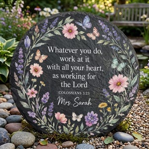 May include: A round black slate garden stone featuring a floral wreath with pink and purple flowers and butterflies. The text reads: Whatever you do, work at it with all your heart, as working for the Lord, Colossians 3:23, and Mrs. Sarah.