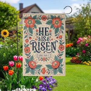 以下が含まれることがあります： ネイビーブルーの筆記体で「HE IS RISEN」と書かれたガーデンフラッグ。オレンジとティールの花の模様で囲まれています。庭と花を背景に、屋外に飾られています。