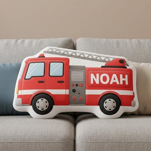 May include: A red and white fire truck shaped pillow with the name "NOAH" printed on the side. The pillow has black wheels and a white ladder on top. The pillow is on a gray couch.