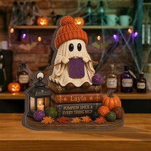 Könnte beinhalten: Eine dekorative Geisterfigur mit orangefarbener Strickmütze, die ein violettes Quadrat strickt und auf einem Stapel Bücher mit der Aufschrift Layla und Pumpkin Spice and Every Thing Nice sitzt. Daneben steht eine schwarze Laterne mit Kerze.