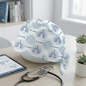 May include: A light blue medical scrub cap with a pattern of lungs and trees, with the name "Dr. DAVID" printed on it. A stethoscope and tablet are in the foreground. The cap is tied in a bow.