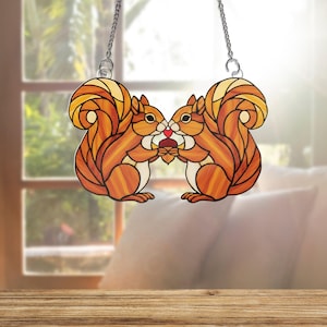 May include: Stained glass window hanging featuring two orange squirrels holding a nut, with a red heart between them. The squirrels have detailed fur and are suspended by a silver chain. The background is a blurred view of a window and a wooden table.