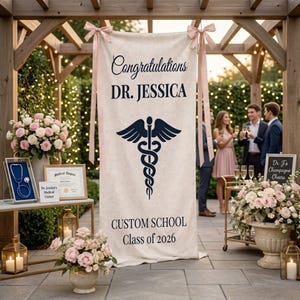 May include: A cream-colored banner with the text "Congratulations DR. JESSICA" and a medical symbol. Below, it reads "CUSTOM SCHOOL Class of 2026." The banner is decorated with pink ribbons and is part of a graduation celebration.