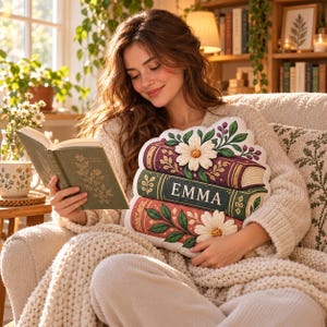 May include: A decorative throw pillow shaped like a stack of three books with floral accents and the name EMMA printed on the center spine. The pillow features a cream background with illustrations of white daisies, green leaves, and purple berries.