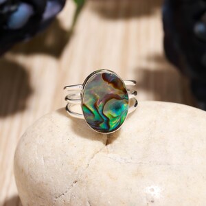 May include: A silver ring with an oval abalone shell center stone. The iridescent shell features swirls of green, blue, and brown. The ring has a triple band design, adding a modern touch to the jewelry piece. The ring is displayed on a white stone.