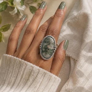 May include: A silver ring featuring a large, oval-shaped moss agate stone with green and grey inclusions. The stone is set in a smooth silver bezel with decorative silver bead accents on the sides of the band, creating a detailed metal frame.