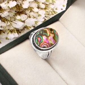 Puede incluir: Anillo de plata con una concha de abulón ovalada, que muestra tonos iridiscentes rosas, verdes y dorados. El anillo tiene una banda de plata detallada y un engaste de bisel. El anillo se muestra en una caja de joyería.