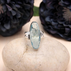 Peut inclure: Une bague en argent ornée d'une grande pierre précieuse ovale bleu clair. La bague est posée sur une pierre de couleur claire, avec des fleurs violet foncé en arrière-plan. La pierre précieuse est facettée, reflétant la lumière.