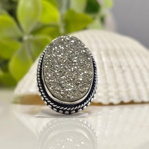 May include: Silver ring with an oval pyrite stone, showcasing a textured, glittering surface. The stone is framed by a detailed, rope-style border. The ring's design is elegant and suitable for everyday wear or special occasions.