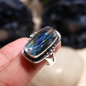 May include: A silver ring featuring a large, rectangular gemstone with iridescent rainbow colors. The gemstone is set in a silver bezel with decorative accents. The ring's band is a simple, polished silver.