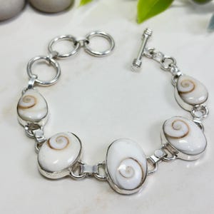 Puede incluir: Una pulsera de eslabones de plata con cinco cabujones ovalados de concha shiva blanca con patrones de espiral marrones. La pulsera incluye un cierre de barra y varios eslabones circulares de plata, con un acabado metálico pulido en todo el diseño.