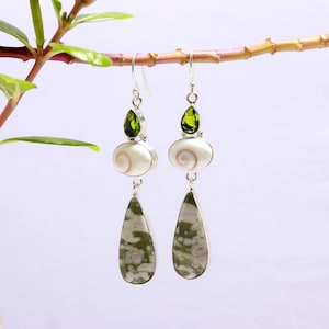 May include: A pair of silver dangle earrings. Each earring has a green gemstone, a white oval shell, and a green and white teardrop-shaped stone. The earrings are suspended from a silver hook. The earrings are a unique design.