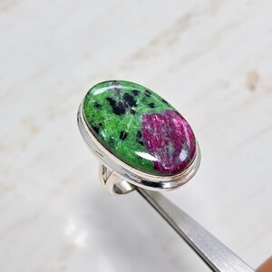 May include: An oval-shaped ring featuring a polished zoisite stone with vibrant green, black, and deep magenta-red inclusions. The gemstone is set in a smooth, polished silver-toned metal band that encircles the base of the stone setting.
