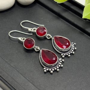 May include: A pair of silver drop earrings featuring a round faceted red gemstone at the top and a larger pear-shaped faceted red gemstone at the bottom. The settings are detailed with small silver beads along the edges and include hook wires.