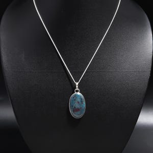 May include: A silver necklace with an oval pendant. The pendant has a blue and red stone set in a silver bezel. The necklace is displayed on a black mannequin.