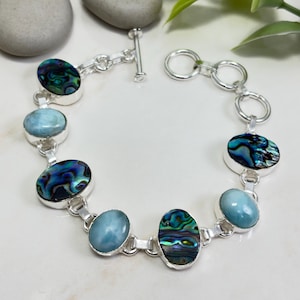 May include: A silver toggle clasp bracelet featuring alternating oval-shaped cabochons of iridescent abalone shell with swirling blue and green patterns and smooth, opaque light blue stones set in polished silver metal settings.