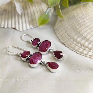 Puede incluir: Un par de pendientes de plata con tres piedras preciosas de color rubí. Los pendientes tienen un diseño de gancho y están colocados sobre un fondo blanco con una concha y vegetación.