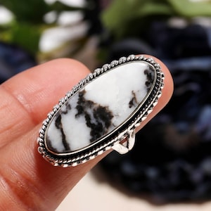 Puede incluir: Anillo ovalado con una piedra blanca con vetas negras, engastado en metal plateado. El anillo tiene un borde texturizado en forma de cuerda. Una joya.