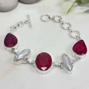 May include: A silver bracelet featuring three faceted, deep red ruby stones and two elongated, irregular white pearls. The stones are set in silver bezels and connected by silver links, with a toggle clasp closure at one end of the chain.