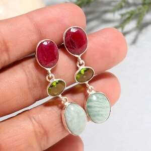 Puede incluir: Pendientes colgantes de plata con piedras preciosas ovaladas. Los pendientes muestran una vibrante combinación de piedras rojas, verdes y celeste. Las piedras están engastadas en una montura de metal plateado.