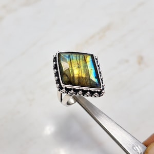 May include: A sterling silver ring featuring a large, kite-shaped labradorite gemstone with iridescent yellow and blue flashes. The stone is set in a bezel with a decorative beaded border, and the band is made of polished silver metal.