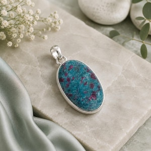 May include: An oval pendant featuring a polished teal-coloured stone with embedded deep magenta inclusions, set in a smooth silver-toned metal frame with a small circular bail at the top for attachment to a chain or cord.