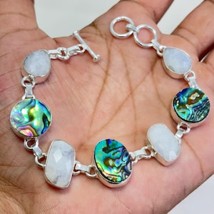 Può includere: Un bracciale a maglie in argento con pietre alternate rotonde e ovali. Il bracciale presenta cabochon in conchiglia di abalone iridescente con motivi vorticosi verdi e blu e gemme bianche traslucide sfaccettate simili alla pietra di luna.