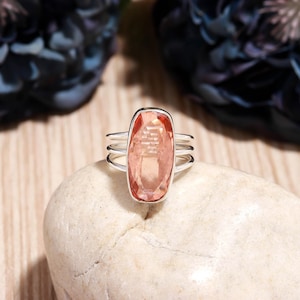 May include: A silver ring with a large, rectangular, peach-colored gemstone. The ring has a triple-band design and is set on a light-colored stone. The background features a wooden surface and dark blue floral accents.