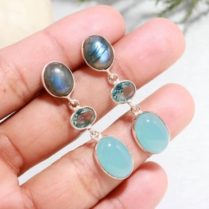 Puede incluir: Un par de pendientes de plata con un diseño colgante. Cada pendiente presenta tres piedras preciosas: una labradorita ovalada en la parte superior, una piedra azul-verdosa ovalada en el medio y una piedra azul claro ovalada en la parte inferior. Los pendientes se sostienen en una mano.