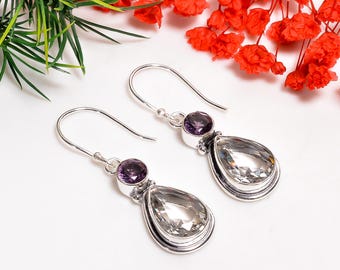 May include: A pair of silver drop earrings featuring a round purple gemstone above a pear-shaped clear gemstone. The earrings are displayed on a white surface with red flowers and green foliage in the background.