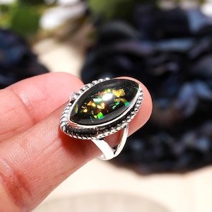 Puede incluir: Anillo de plata con una piedra preciosa negra ovalada con inclusiones iridiscentes doradas y verdes. La piedra preciosa está engastada en un bisel de plata decorativo con un diseño de cuerda trenzada. La banda del anillo es de plata pulida.