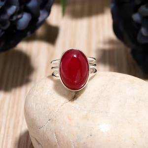 以下が含まれることがあります： 大きな楕円形の深い赤色の宝石が付いたシルバーリング。リングはトリプルバンドのデザインで、明るい色の丸い石の上にセットされています。背景には、濃い紫色の花のアクセントと木の表面があります。