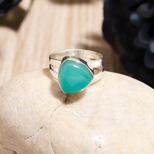 May include: A silver ring with a split band design, featuring a triangular, light teal-colored gemstone. The ring is set on a light-colored stone, showcasing the jewelry's details.